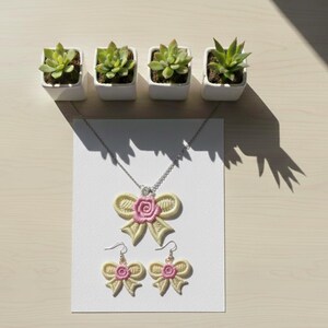 May include: A necklace and earrings set featuring a bow design with a pink rose center. The jewelry is cream-colored with pink accents and is displayed on a white card. Four small potted succulents are arranged above the jewelry.