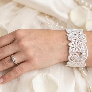 Può includere: Una mano con un anello di fidanzamento in argento e un braccialetto in pizzo bianco con un motivo floreale. Gli oggetti sono su uno sfondo bianco con perle e petali di fiori.