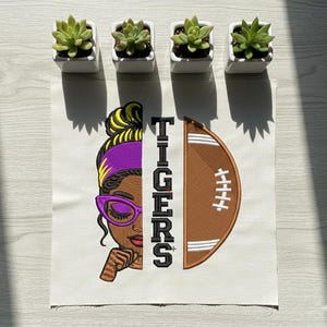 May include: Embroidered design featuring a woman with purple glasses and a headband, a football, and the word "TIGERS" in black. The design is on a white fabric. Four small potted succulents are arranged above the design.