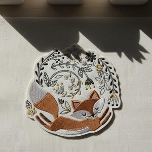May include: Circular embroidered patch depicting a sleeping fox in brown, grey, and white, encircled by a floral wreath with black, gold, and white details. The patch showcases a detailed design.