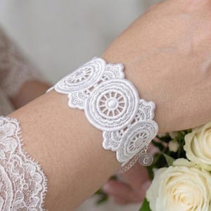 May include: A white lace bracelet with a circular design is worn on a wrist. The bracelet is made of delicate lace and features a series of connected circular patterns. The wrist is next to a bouquet of white roses and greenery. The sleeve of a white lace dress is visible.
