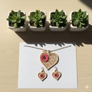 May include: A handmade jewelry set featuring a heart-shaped pendant necklace and matching earrings. The pendant and earrings are beige with a pink button accent. The set is displayed on a white card, with four small potted succulents in the background.