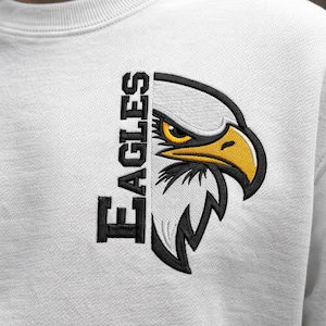 Puede incluir: Sudadera blanca con un diseño de cabeza de águila bordada. La cabeza del águila está dividida verticalmente, con la palabra "EAGLES" en el lado izquierdo. El águila tiene un pico y un ojo amarillos, con detalles en blanco y negro.