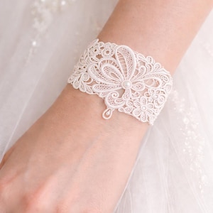 May include: Ivory lace cuff bracelet with floral and teardrop details. The delicate design features a central flower with a pearl accent. This elegant accessory is perfect for weddings and special occasions.