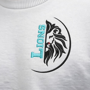 May include: White sweatshirt with an embroidered design featuring a lion's head and the word "LIONS" in turquoise. The lion's head is black and detailed, set within a black oval.