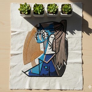 May include: An embroidered patch featuring a cubist-style portrait in shades of blue, brown, and black, on a white fabric. Four small white square pots with green succulents are arranged above the patch.