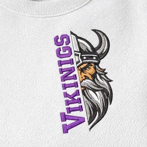 May include: White sweatshirt featuring a Viking graphic. The design includes a detailed illustration of a Viking with a horned helmet and a long beard, with the word "VIKINGS" in purple vertically on the left side.