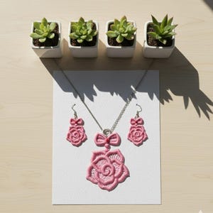 Puede incluir: Un conjunto de joyas bordadas en rosa con un collar con colgante de rosa y pendientes a juego. El conjunto se exhibe sobre una tarjeta blanca, con pequeñas suculentas en macetas blancas en el fondo. La joyería tiene un diseño delicado y femenino.