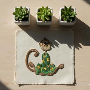 May include: A decorative embroidered patch featuring a cartoon monkey in a green outfit with yellow accents. The patch is on a cream-colored fabric. Three small square white planters with green succulents are arranged above the patch on a light wood surface.