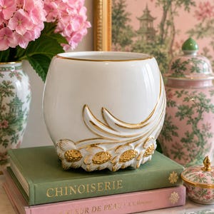 May include: A white ceramic vase with a rounded shape and gold-painted relief details featuring stylized floral motifs around the base. The rim of the vase is also accented with a thin gold line, creating a polished finish on the smooth white surface.