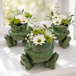 May include: Three glossy green ceramic frog-shaped planters, each featuring a sculpted frog base and a wide opening. The planters are filled with arrangements of white daisies, small green chrysanthemums, and delicate white baby's breath flowers.