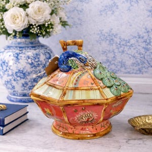 May include: A decorative ceramic peacock-themed container with a lid. The container is primarily red with gold trim and a handle. The lid features a peacock head and tail. A blue and white floral vase is in the background.