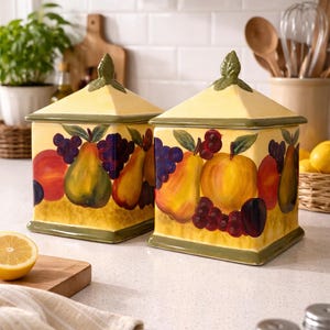 May include: Two yellow ceramic canisters with lids, decorated with painted fruit including pears, grapes, and peaches. The lids are topped with a green leaf design. The canisters are square and sit on a white countertop.