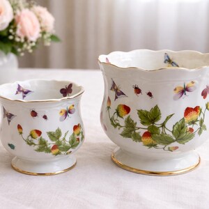 May include: Two white ceramic planters with scalloped rims and gold trim. The planters are decorated with colorful butterflies, ladybugs, and strawberries with green leaves. The planters are on a white surface.