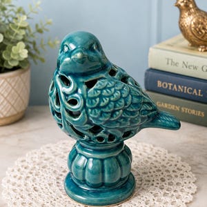 May include: A teal ceramic bird figurine featuring intricate cut-out patterns on its body and wings. The bird is perched on a rounded, fluted pedestal base. The glossy glaze finish reflects light, highlighting the detailed sculpted feathers and eyes.