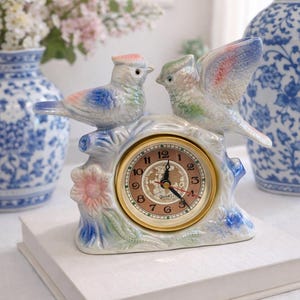 May include: A ceramic bird-shaped clock with a gold-tone clock face. The clock features two birds perched on a branch with a pink flower. The birds have blue, pink, and green accents. The clock sits on a white surface.