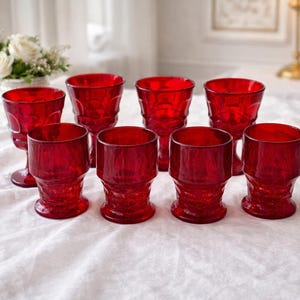 May include: A set of eight ruby red drinking glasses. The glasses have a textured, geometric design and are arranged on a white tablecloth. A vase of white flowers is in the background.