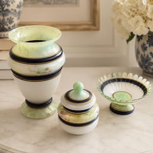 May include: A collection of decorative glass objects, including a vase, a small lidded jar, and a pedestal bowl. The objects feature white, green, and navy blue accents with gold trim. The vase is the tallest, with a flared top.