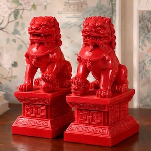 May include: Two matching red Foo Dog statues. Each statue is in a seated position atop a square pedestal. The Foo Dogs have detailed manes, and are holding a ball under their paws. The pedestals have intricate carvings.