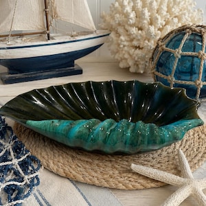 May include: A ceramic bowl shaped like a large seashell with a glossy finish. The interior features a deep dark green color, while the exterior rim transitions into a vibrant turquoise blue. The bowl has a fluted, scalloped edge design.