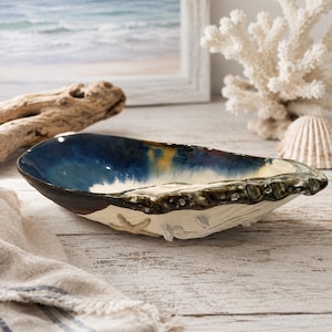 May include: A ceramic bowl shaped like an oyster shell with a glossy interior featuring deep blue, yellow, and white glaze. The exterior has a textured, matte off-white finish with sculpted shell details and a dark, mottled brown rim.