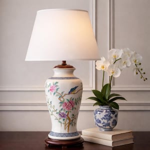 May include: A decorative table lamp with a white lampshade and a ceramic base. The base features a floral and bird design in pink, purple, and blue. The lamp sits on a dark wooden base, with a white orchid and a blue and white vase nearby.