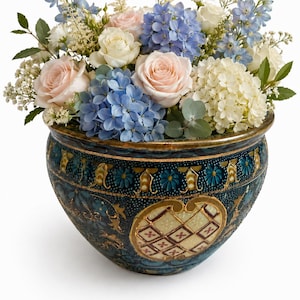 May include: A floral arrangement featuring light pink roses, white roses, blue hydrangeas, white hydrangeas, and blue delphiniums in a dark blue ceramic bowl with gold-toned decorative relief patterns and a central cream-colored diamond grid panel.