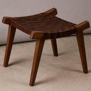 May include: A small, handcrafted wooden stool with a woven leather seat. The stool features four angled legs and a dark brown finish. The seat is made of interwoven strips of brown leather. The design is minimalist and modern.