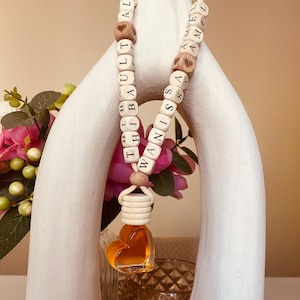 Peut inclure: Un désodorisant pour voiture avec un flacon en verre en forme de cœur rempli de liquide ambré. Le désodorisant est suspendu à un collier de perles avec des perles de lettres en bois épelant des noms.