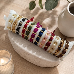 Peut inclure: Une collection de onze bracelets disposés sur un coussin de présentation cylindrique blanc. Chaque bracelet présente un design de maillons en métal doré entrelacés avec des rubans en tissu de diverses couleurs, dont crème, noir, rouge, vert et rose.