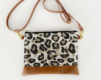 May include: A crossbody bag with a leopard print fabric upper and a brown leather bottom. The bag has a brown leather strap and gold-colored hardware. The bag is rectangular and has a zipper closure.