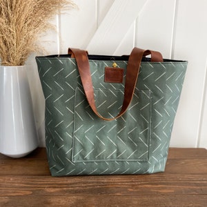 May include: A green canvas tote bag with white diagonal lines and brown leather straps. The bag has a small pocket on the front and a leather tag that says "Something Nice".