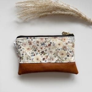 May include: A small, rectangular pouch with a floral pattern on the top half and brown leather on the bottom. The floral pattern features cream, brown, and blue flowers on a white background. A gold zipper and the text "SEPTEMBER SKYE" are visible.