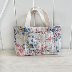 Puede incluir: Un bolso tote acolchado con un diseño de patchwork con estampados florales en rosa, azul y amarillo. El bolso tiene un pequeño bolsillo frontal y asas de color crema. La etiqueta del bolsillo dice "SEPTEMBER SKYE".