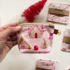 Puede incluir: Un monedero rosa con cremallera y un estampado de casa de jengibre, con herrajes dorados y una lengüeta de cuero marrón. Cerca hay un pequeño árbol de Navidad rojo y varios marcapáginas de tela a juego.