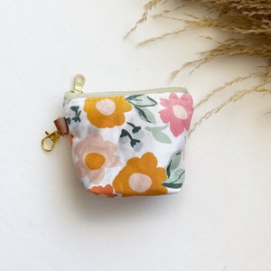 Puede incluir: Una pequeña bolsa blanca con cremallera con estampado floral. La bolsa tiene una cremallera dorada y una correa de cuero marrón con un cierre dorado.