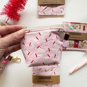 Puede incluir: Una bolsa rosa con cremallera con un patrón de Papá Noel blanco y rojo. La bolsa está hecha de tela y tiene una cremallera dorada. La bolsa está sobre una superficie blanca. El texto "BOOKMARK" está en una etiqueta marrón unida a la bolsa.
