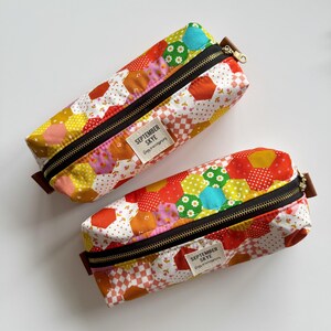May include: Two colorful pencil cases with a patchwork design. The cases feature a variety of hexagonal fabric pieces in red, yellow, orange, and green, with a black zipper and a small tag that reads "SEPTEMBER SKYE".
