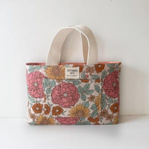 Puede incluir: Una pequeña bolsa de mano floral con asas de lona blanca. La bolsa está hecha de una tela de color crema con un estampado floral en tonos rosa, naranja y verde. La bolsa tiene un pequeño bolsillo en la parte delantera con el texto "SEPTEMBER SKYE" impreso en él.