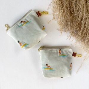 Puede incluir: Dos pequeñas bolsas con cremallera de color claro con un patrón repetido de surfistas en tablas de surf amarillas. Cada bolsa tiene una cremallera dorada, una lengüeta de cuero marrón y un cierre dorado. Las bolsas están sobre una superficie blanca con plantas secas.
