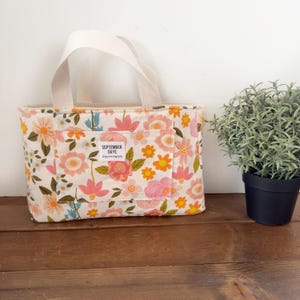 May include: A white tote bag with a floral print featuring pink, orange, and yellow flowers. The bag has a white handle and a small pocket on the front with the text "SEPTEMBER SKYE" and "happy happenings".