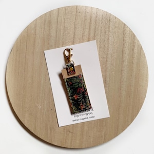 May include: A small, green and pink floral patterned leather chapstick holder with a gold metal keychain attached. The holder is on a white card with the text "SEPTEMBER SKYE bags & accessories leather chapstick holder".