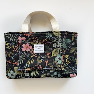 May include: Black tote bag with a floral print featuring pink, green, and yellow flowers. The bag has a white handle and a label that reads "SEPTEMBER SKYE".