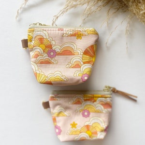 May include: Two pink zippered pouches with a retro 70s style sun and cloud pattern. The pouches are different sizes.