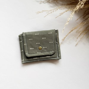 May include: Olive green fabric wallet with a white dotted pattern and a brass snap closure. The wallet is rectangular with a flap and white stitching around the edges. A simple, stylish accessory.