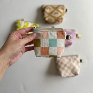 Puede incluir: Una pequeña bolsa con cremallera con un patrón a cuadros en tonos rosa, verde y marrón. La bolsa está hecha de tela y tiene una cremallera dorada.