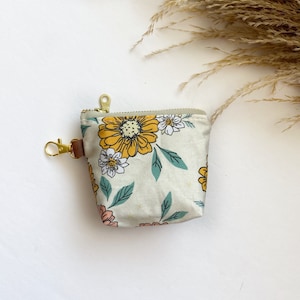May include: A small, beige zippered pouch with a floral print. The print features yellow, pink, and white flowers with green leaves on a beige background. The pouch has a gold zipper and a gold key ring.
