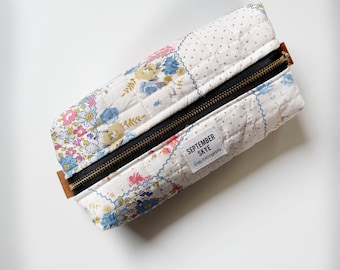 Puede incluir: Estuche acolchado con estampado floral en tonos rosa, azul y amarillo. El estuche tiene una cremallera negra y una pequeña etiqueta que dice "SEPTEMBER SKYE bags & accessories."