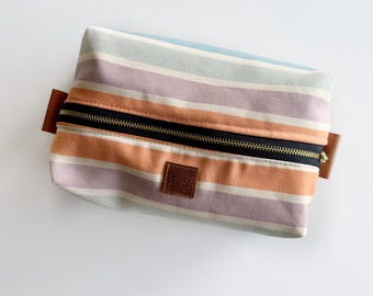 May include: A striped canvas makeup bag with a zipper closure and a brown leather patch with the initials "SS" on it.