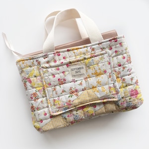 Può includere: Una borsa tote trapuntata con un motivo floreale in rosa, giallo e arancione su sfondo bianco. La borsa ha una tasca frontale e un'etichetta con la scritta "SEPTEMBER SKYE bags & accessories". Ha un manico color crema.